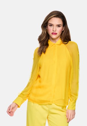 Woman wearing a yellow sheer long-sleeve blouse and matching yellow pants, posing with a neutral expression on a white background.