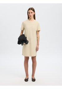Bežinė trumpomis rankovėmis T-shirt suknelė su tiesiu siluetu, derinama su juoda pintine rankine, užsidėta su juodais plokščiais batais. Sklandi tekstūra, minimalus dizainas.