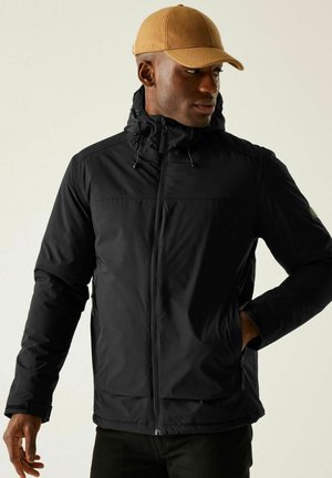 Mężczyzna w beżowej czapce i czarnej kurtce z kapturem, z rękami w kieszeniach, patrzący w prawo na jednolitym jasnym tle.