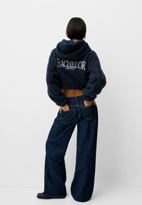 Mörkblå, kortärmad, plysch-hoodie med "BACHELOR" broderat på baksidan, tillsammans med vida mörka denimjeans.