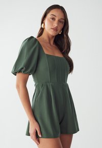 Combi-short verte à manches ballon avec un décolleté carré, des plis sur le devant et un corsage ajusté. Tissu lisse ; longueur short. Boucles d'oreilles en or en forme de cercle pour accentuer.