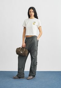 Λευκό βαμβακερό t-shirt με κεντήματα λουλουδιών, συνδυασμένο με φαρδιά γκρι παντελόνια cargo και μια σκούρα καφέ τσάντα. Το σύνολο ολοκληρώνουν γκρι αθλητικά παπούτσια.