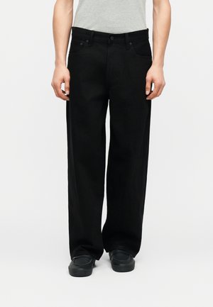 Inimene, kes kannab lahtisi musti pükse ja musti kingi, seisab käed rüpes valge lihtsa tausta ees.