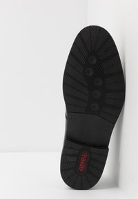 Suola di scarpa in gomma nera con un motivo testurizzato, caratterizzata da un design circolare e un piccolo logo di marca rosso. Forma piatta e arrotondata.