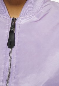 Fialová bomber bunda s hladkou, lesklou texturou, ribbovaným límcem a stříbrným zipem s černým táhlem, která se vyznačuje minimalistickým designem.