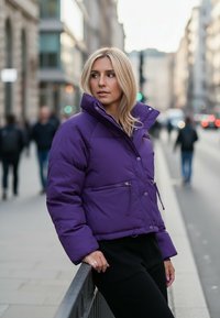 Fioletowa kurtka puchowa z wysokim kołnierzem, kieszeniami zapinanymi na suwak i guzikiem z przodu. Materiał wydaje się błyszczący, na tle miejskiej ulicy.