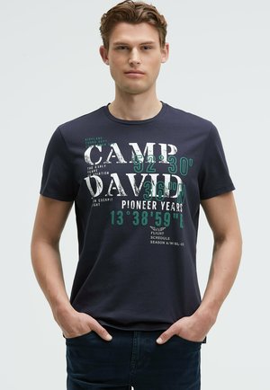 Junger Mann trägt ein dunkelblaues Camp David T-Shirt mit weißem und grünem aufgedrucktem Text und Koordinaten, Hände in den Taschen, steht vor einem schlichten Hintergrund.