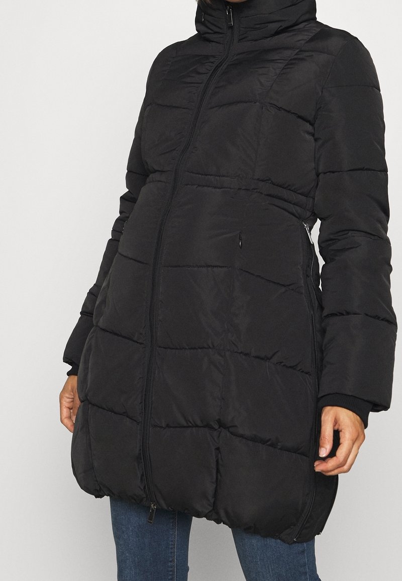 Personne portant un long manteau matelassé noir avec un col montant et une fermeture éclair à l'avant, les mains détendues le long du corps, associé à un jean bleu.