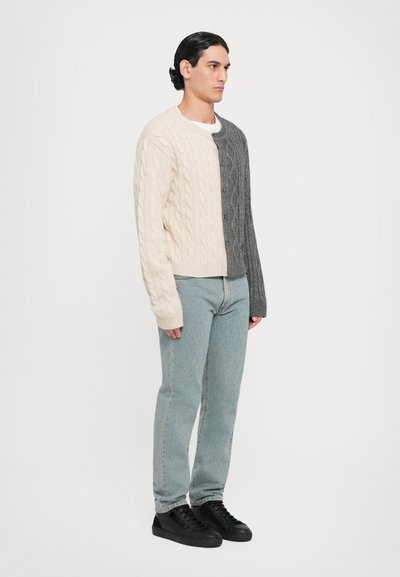 Stickad cardigan med en delad design i ljus beige och mörkgrå, med ett texturerat kabelmönster och framknappar. Kombinerad med ljusblå jeans och svarta sneakers.