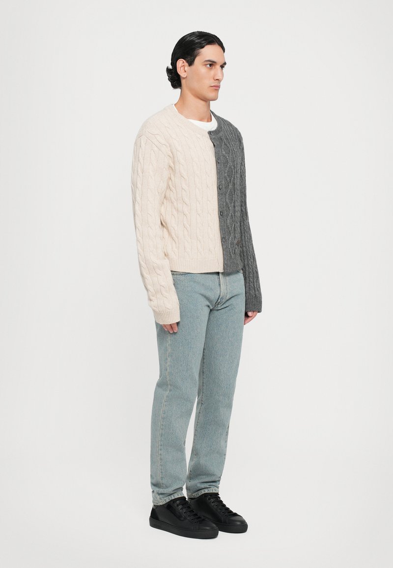Cárdigan de punto con un diseño dividido en beige claro y gris oscuro, con un patrón de trenzado texturizado y botones en el frente. Combinado con jeans azul claro y zapatillas negras.