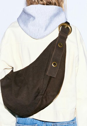 Személy világos dzsekit és szürke kapucnis pulóvert visel, és egy nagy sötétbarna bőr oldaltáska van nála, sárgaréz csattal és szegecsekkel.