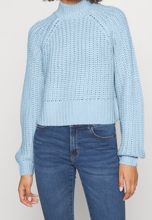 Personne portant un pull en grosse maille bleu clair à manches longues et col montant associé à un jean ajusté bleu foncé.