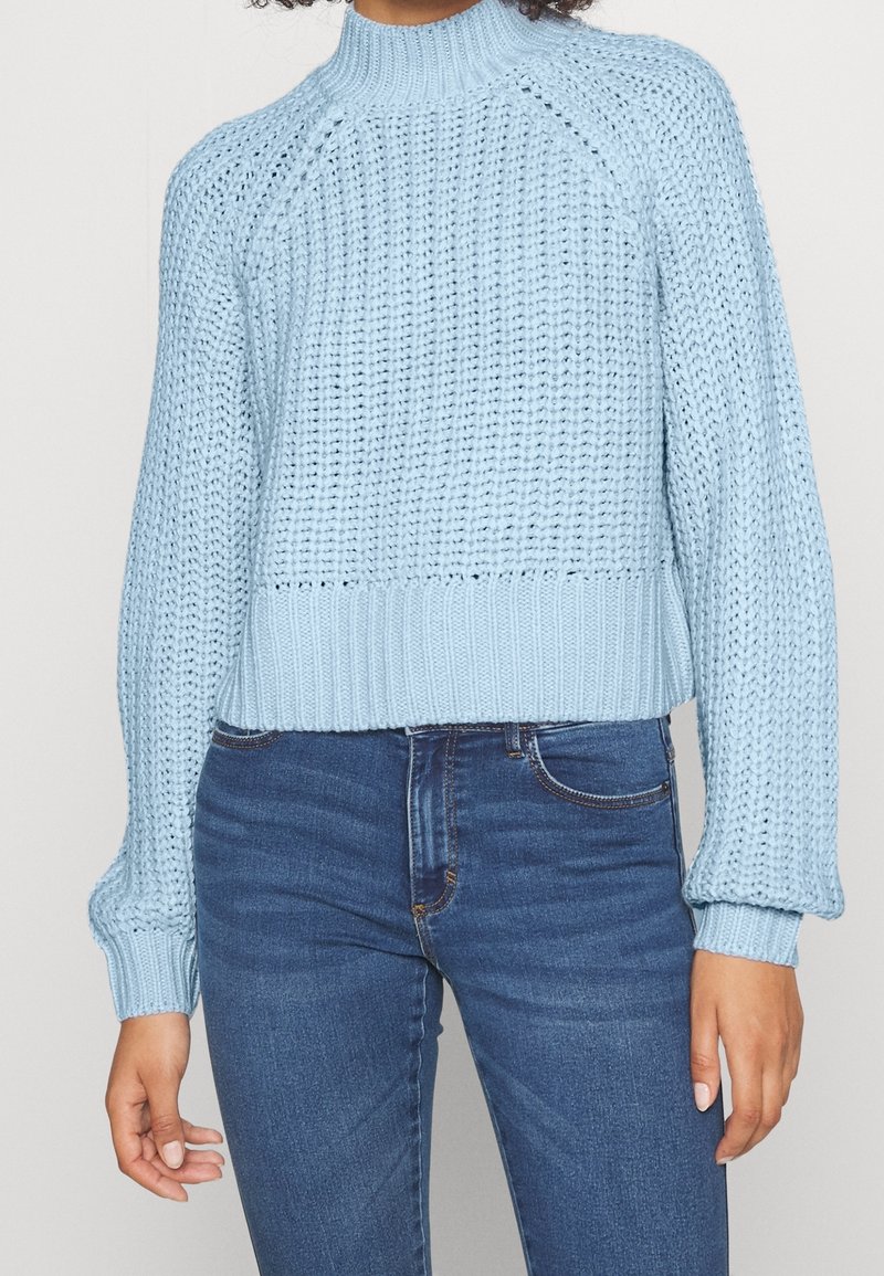 Personne portant un pull en grosse maille bleu clair à manches longues et col montant associé à un jean ajusté bleu foncé.