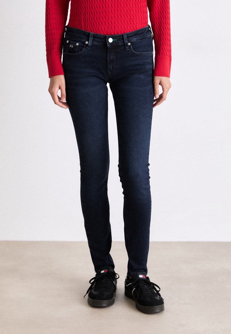 Mujer vestida con jeans ajustados de color azul oscuro, zapatillas negras con logotipos y un suéter de punto en color rojo, de pie sobre un suelo claro contra una pared blanca.
