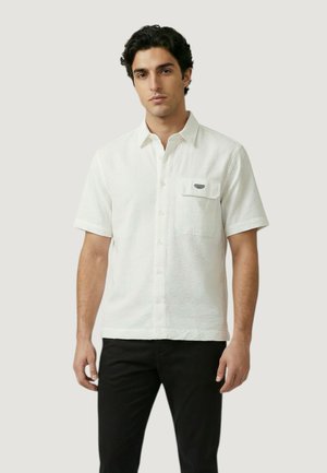 Hombre con cabello oscuro rizado que lleva una camisa blanca de manga corta con textura y botones, y pantalones negros, de pie contra un fondo claro y liso.