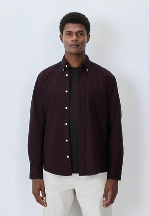 Dunkellila Button-up-Hemd mit weißen Knöpfen und einer Brusttasche, über einem schwarzen T-Shirt getragen. Glatte Textur, lange Ärmel.