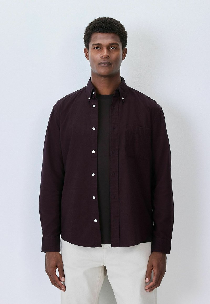 Dunkellila Button-up-Hemd mit weißen Knöpfen und einer Brusttasche, über einem schwarzen T-Shirt getragen. Glatte Textur, lange Ärmel.