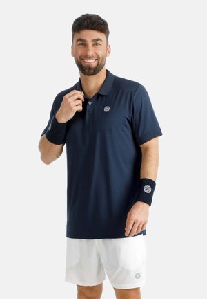 Hombre sonriente con polo azul marino, pantalones cortos blancos y muñequeras a juego, de pie frente a un fondo claro y liso.