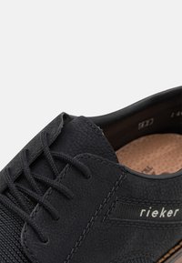 Chaussure noire à lacets en cuir texturé, avec un panneau latéral en maille, des coutures contrastées et une semelle marron. Marquée du label "rieker".