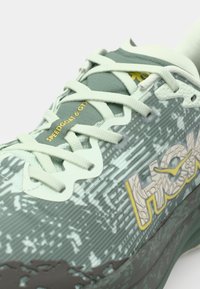 Chaussure de course verte avec un dessus en mesh léger, lacets blancs et semelle en caoutchouc. Présente un logo jaune et des accents texturés.