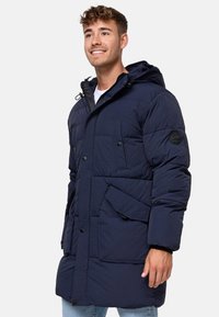 Jachetă puffer bleu închis cu glugă, închidere cu butoane și fermoar, multiple buzunare și un finisaj texturat. Include un patch cu logo pe mânecă.