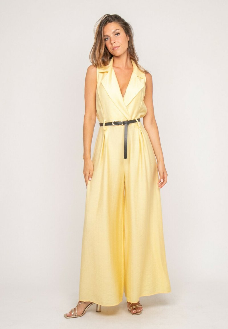 Femme portant une combinaison jaune sans manches à jambes larges avec un décolleté en V profond et une ceinture noire, debout devant un fond uni.