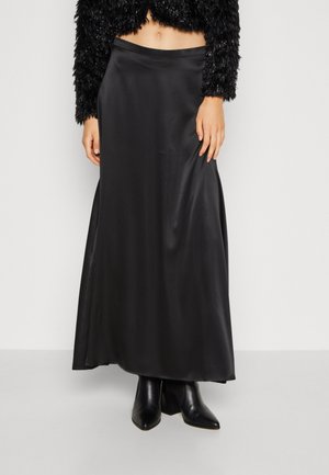 Vrouw die een zwarte satijnen maxi-rok draagt, zwarte enkellaarsjes met puntige neus en een cropped, getextureerde zwarte top met lange mouwen, staand tegen een witte muur.
