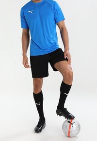 Blaue Sportshirt mit kurzen Ärmeln, schwarze Shorts, schwarze Socken und schwarze Fußballschuhe. Ein silberner Fußball mit schwarzen Akzenten liegt zu Füßen.
