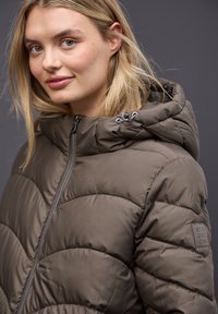 Chaqueta de plumas marrón con cierre de cremallera, que presenta una capucha y un diseño acolchado. Parche con el logotipo de la marca visible en el hombro. Textura suave.