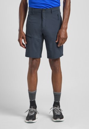 Marineblaue Shorts aus leichtem Stoff, mit einer Reißverschlusstasche, Gürtelschlaufen und einer taillierten Passform. Getragen mit schwarzen Turnschuhen und grauen Socken.