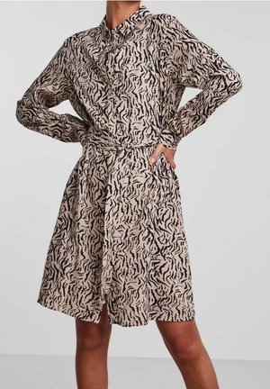 Kobieta w beżowej sukience koszulowej z długim rękawem i czarnymi, abstrakcyjnymi paskami w stylu tygrysa, stojąca z rękami na biodrach na jednolitym tle.