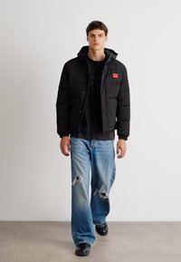 Giubbotto imbottito nero con cappuccio, dotato di una toppa rossa con logo, abbinato a jeans chiari strappati e scarpe nere.