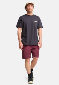 Mørkegrå bomulls t-skjorte med en liten logo, sammen med burgunder shorts og svarte joggesko med hvite såler og mørke sokker.