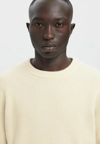 Kreemjas soonikuga kampsun ümmarguse kaelusega ja tekstuurse kangaga, millel on peen muster ja kaeluse ümber õmmeldud detailid.