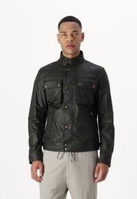 Black leather jacket with high collar, two chest pockets, zipper pockets, and brass hardware accents. Worn over light gray trousers.