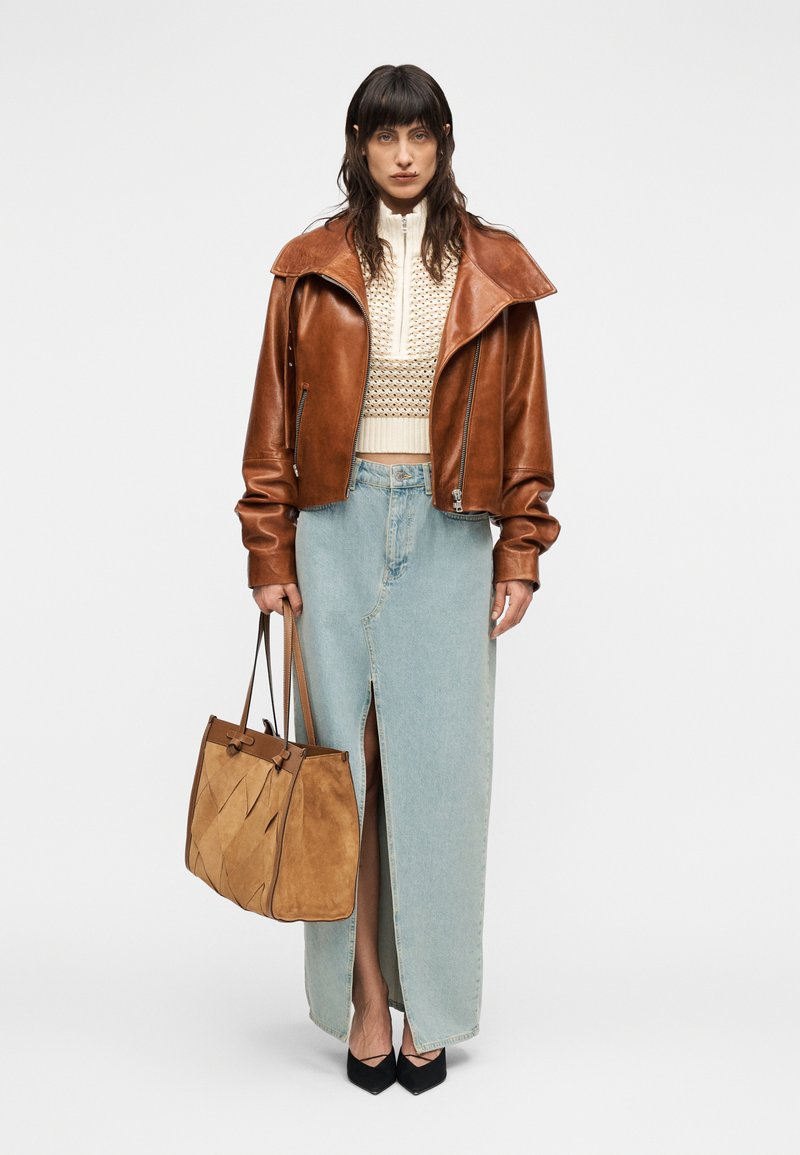 Veste courte en cuir marron, haut en maille crème, jupe longue en denim bleu clair avec une fente avant, sac en daim marron et escarpins noirs à bouts pointus.