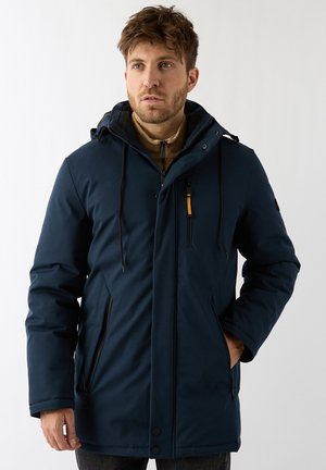 Man met kort bruin haar draagt een donkerblauwe winterparka met capuchon, ritszakken en een bruine binnenlaag, staande tegen een effen achtergrond.