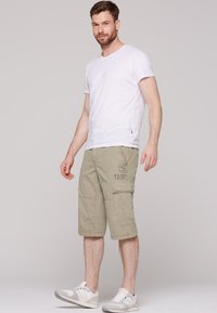 Biela tričko s okrúhlym výstrihom v kombinácii s khaki cargo šortkami po kolená, ktoré majú vrecká a potlač. Model má na sebe šedé a biele tenisky.