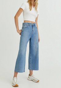 Jeans larges bleu clair à taille haute et design classique à cinq poches, associés à un polo blanc ajusté et des baskets.