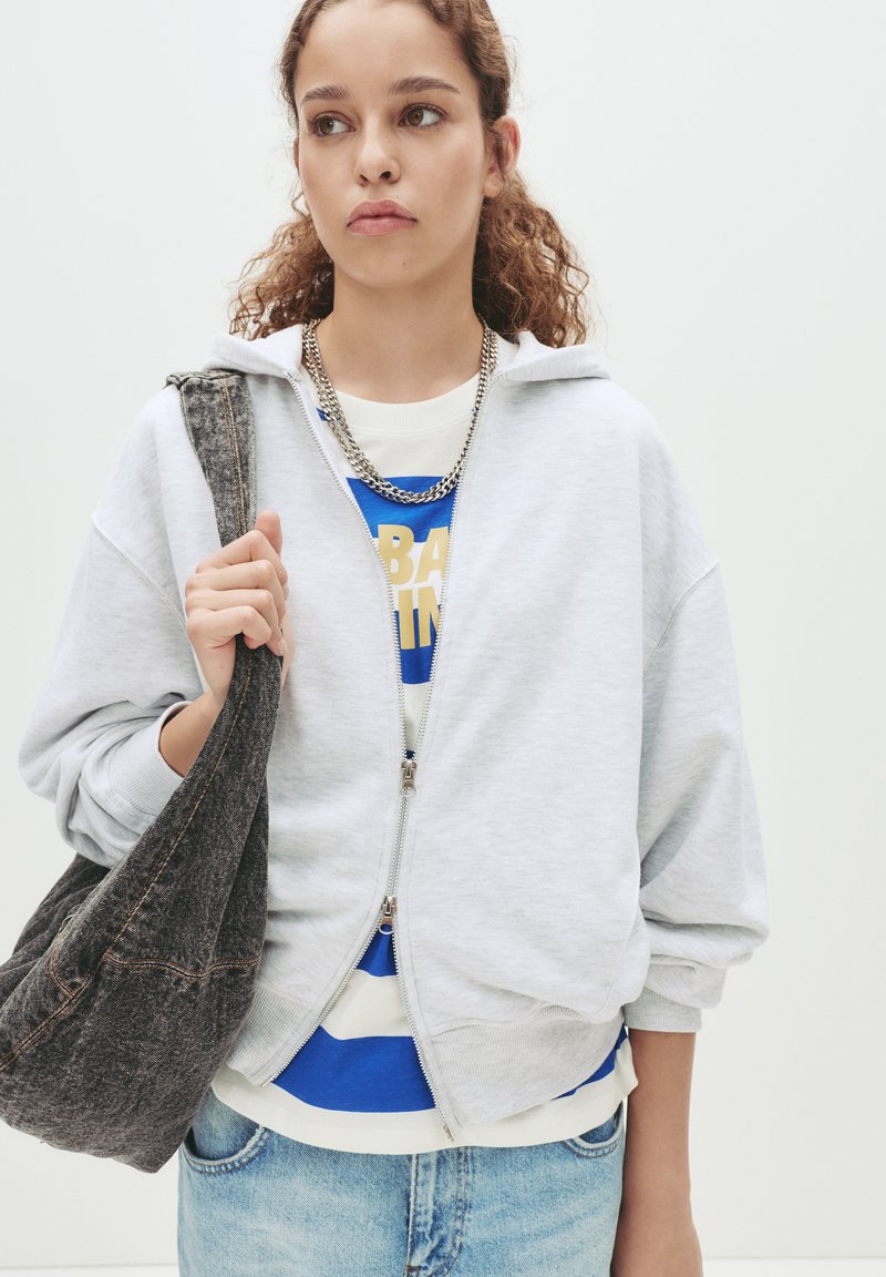 Lys grå zip-up hoodie med en afslappet pasform, båret over en blå og hvid stribet skjorte. Grå denim skuldertaske og sølv kædemand visible.