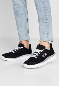 Baskets de sport noires avec un dessus en maille texturée, des lacets blancs et une semelle légère blanche. Présente un détail de logo sur le côté.