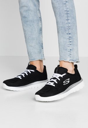 Zapatillas deportivas negras con parte superior de malla texturizada, cordones blancos y suela blanca ligera. Presenta detalles del logo en el lateral.
