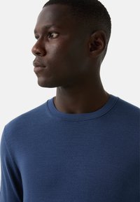 Młody mężczyzna o ciemnej karnacji, ubrany w niebieski sweter z okrągłym dekoltem, patrzy w lewo na jednolity jasny tle.