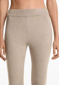 Close-up van een persoon die beige gebreide tights draagt met een geribbelde elastische tailleband, waarbij de handen ontspannen aan de zijkanten hangen.