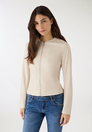 Giovane donna con capelli lunghi e scuri, indossa una giacca beige con cerniera e jeans blu, in piedi contro uno sfondo bianco semplice.