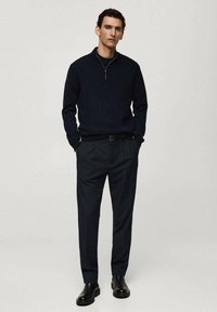 Mornarska zip-up dukserica, črna turtleneck majica pod njo, v kombinaciji s temnimi hlačami in črnimi gležnjači. Minimalističen dizajn s premišljeno silhueto.