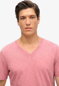 Ljust rosa V-ringad t-shirt gjord av mjukt, strukturerat tyg. Korta ärmar och avslappnad passform, med subtila sömmar runt halsen.