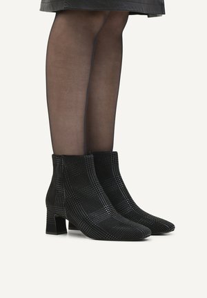 Woman wearing black patterned ankle boots with block heels and sheer black stockings, standing against a plain background.