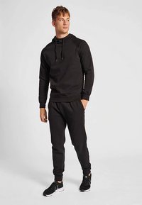 Sort hættetrøje med snore og ribbede ærmeafslutninger, kombineret med matchende sorte joggingbukser. Bruges med sorte atletiksneakers med minimalistisk design.