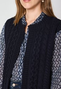 Gilet lavorato a maglia di colore blu navy con motivo a treccia, scollatura rotonda e bordi a costine, indossato sopra una camicetta blu a fantasia.